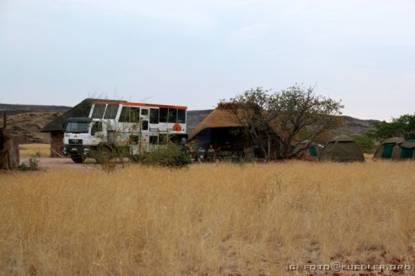 IMG_4168 <P><B>Damara Camp</B></P>

<div align=left>
<P>Eigentlich sollte es jetzt an der Skelettküste entlang in Richtung Norden weitergehen. Aber leider können wir nicht wie geplant unseren Weg fortsetzen, weil die dicht an der Küste
liegende Straße teilweise unterspült ist und vor dem Befahren gewarnt wird. Eine Panne ist auf der Strecke, auf der selten ein Auto vorbeikommt, nicht anzuraten.</P>

<P>Wir nehmen stattdessen einen Weg, der uns direkt an der Spitzkoppe und am Brandberg vorbeiführt. Am Nachmittag erreichen wir unser heutiges Camp mitten zwischen einigen Tafelbergen im Steppengras. Rundherum ist nichts. Das Camp wird von einigen Damara betrieben. Es
gibt eine Bar und erstaunlich saubere Klos und Duschen. Warmwasser machen wir mit einem Ofen, über dem ein großer Kessel hängt.</P>
</div>